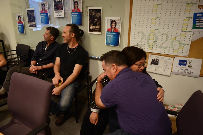 Susan Han hugs City Inside/Out host Brian Callanan following a tribute video he helped create for her on her last day at Seattle Channel. At left are Vincent Pierce and Brett Rudy. Posters of Susan Han hang on the wall.