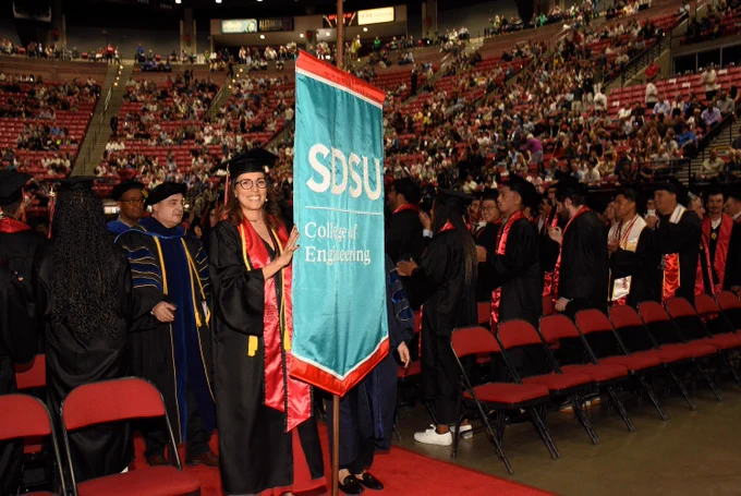 A person in graduation attire holding a banner that reads "SDSU College of Engineering" leads a line of graduates in a ceremony hall filled with seated attendees.