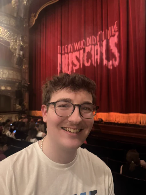 Twitter user @frickpickles smiling in front of the stage curtain for ‘The Guy Who Didn’t Like Musicals’  West End debut at the Apollo Theatre.