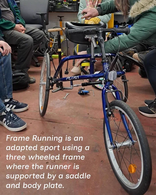 A photo of a blue child frame runner in a workshop with the text stating 'Frame Running is an adapted sport using a 3 wheeled frame where the runner is supported by a saddle and body plate.'