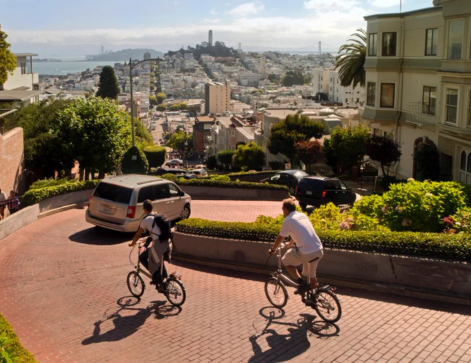 Two cyclists navigate a winding brick road descending a steep hill with a cityscape and bay in the background under a clear sky.