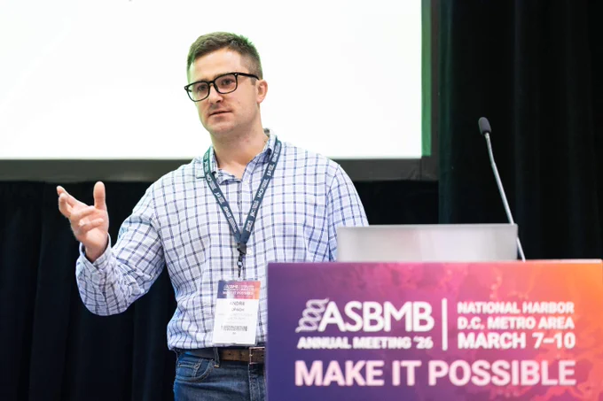 Speaker presenting at the 2026 ASBMB Annual Meeting with a laptop and microphone and large screen in the background.
