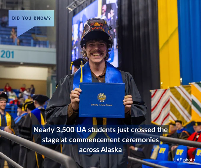 A graduate, smiling widely, holds a blue diploma folder with a gold emblem at a commencement ceremony. The graduate wears a unique hard hat and a blue stole, standing in front of a blurred background of attendees and colorful flags. A text overlay reads, "Nearly 3,500 UA students just crossed the stage at commencement ceremonies across Alaska." A speech bubble in the top left asks, "Did you know?"