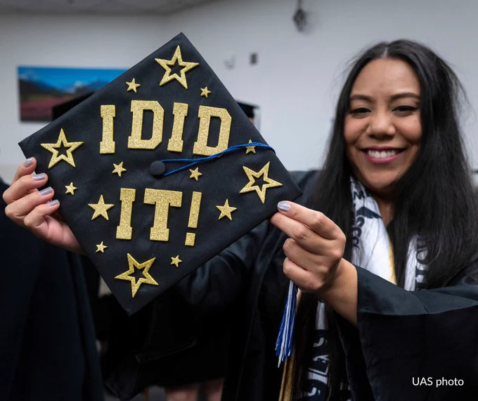 A graduate in a cap and gown smiles while holding up a black graduation cap decorated with glittery gold letters spelling "I DID IT!" and surrounded by star shapes.