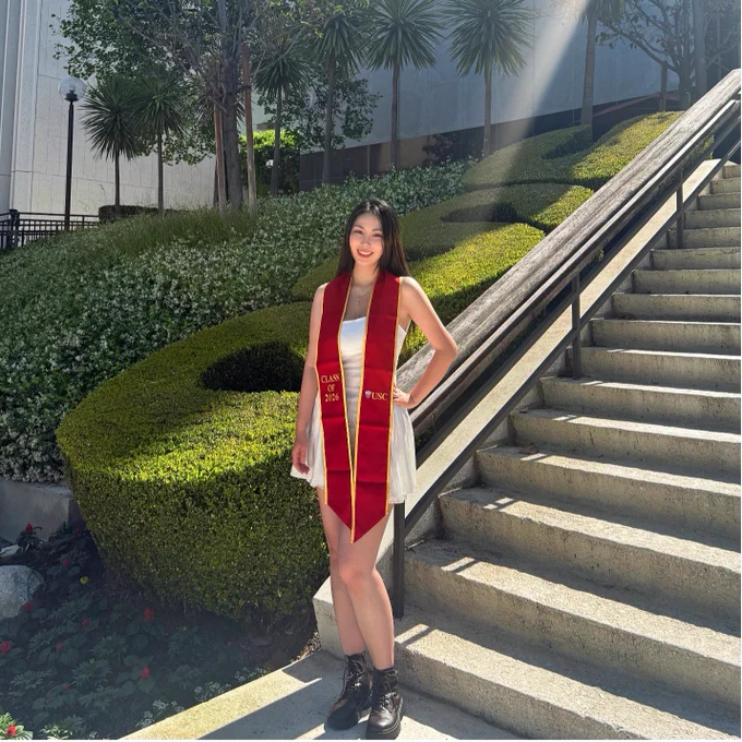 A person stands on outdoor steps next to neatly trimmed greenery, wearing a USC graduation stole. Sunlight filters through overhead trees.