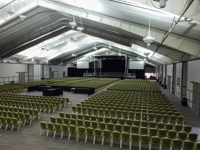 A large, empty Exhibition Hall at French Lick Resort with rows of green chairs facing a stage. The stage features black curtains and two large screens on either side. The space is well-lit, with a high, white ceiling.