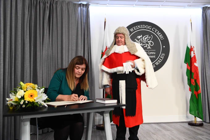Sioned Williams is seated at a table, writing in a book (words not visible). On the table is a flower arrangement of yellow and white flowers to one side of her, and a bible to the other. To Sioned’s right a man in full official outfit in red in stood watching over her shoulder. Behind the man and partially obscured is a large black plaque (official seal) on the wall which has the words “Prif Weinidog Cymru, First Minister of Wales” and an outline of a Welsh dragon in white. Either side of the official seal is a Welsh flag. Black curtains have been drawn to reveal the flags and official seal. Sioned is seated in front of one of the drawn curtains. 