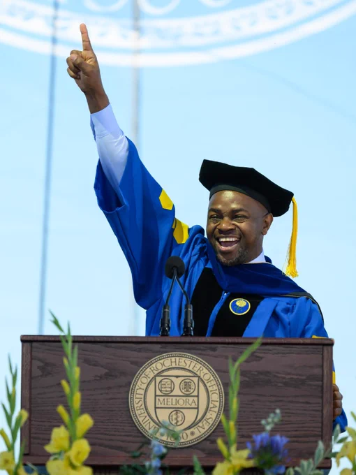 Onesmo Mushi, in graduation cap and gown, points to the sky and smiles from behind the podium at Commencement. 