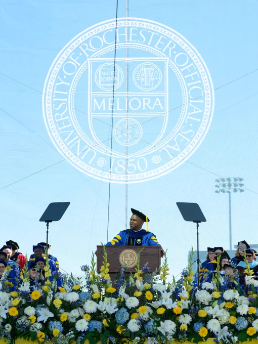 Onesmo Mushi speaking from the stage at Commencement, behind a podium surrounded by flowers. 