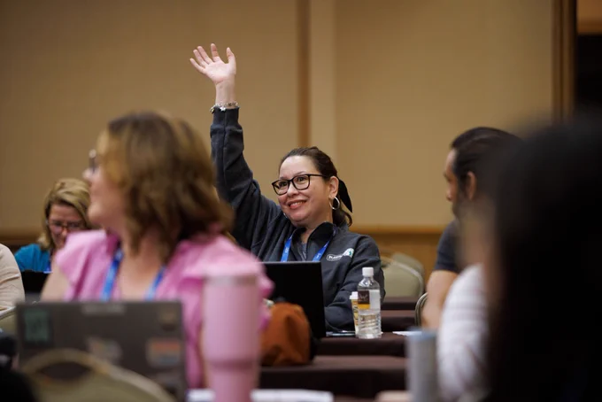 Photo of several people sitting at tables in a room. One person is in focus, and they are smiling while raising their hand to someone off camera. 