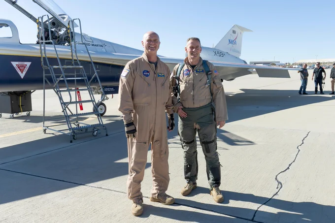 NASA's two X-59 test pilots, wearing tan flight suits, stand on the runway in front of their quiet supersonic X-59 plane. The sky behind them is bright and blue. Credit: NASA/Genaro Vavuris