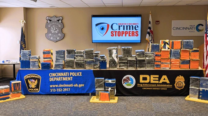 Stacks of confiscated drugs displayed on tables with Cincinnati Police and DEA banners