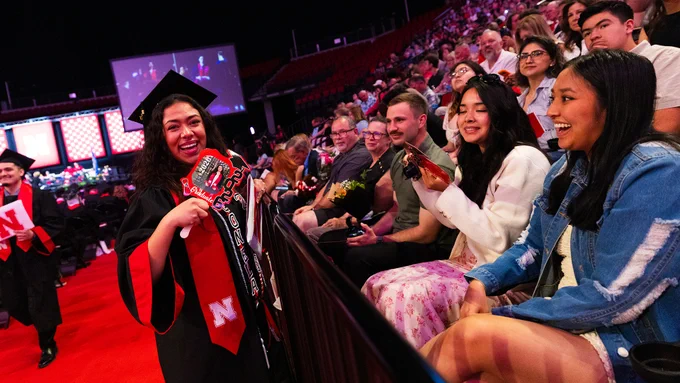 A female graduate wearing a black robe, black mortarboard and red stole smiles as she stands next to her family in the front row of the lower level in Pinnacle Bank Arena.