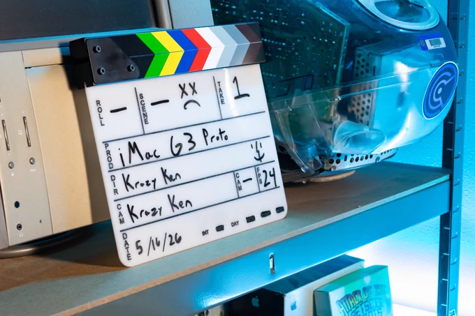 A film slate sitting on a back shelf in Krazy Ken's lair. A 20th Anniversary Macintosh is behind it, and a rare iMac G3 prototype is on the right of it. The prototype has no EMI shielding and the case is smooth and transparent. The slate says, take: 1, production: iMac G3 proto, director: Krazy Ken, date: 5/16/26, interior, day
