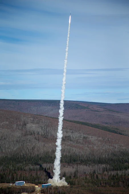 A rocket launches from a facility surrounded by forest, leaving a long trail of white smoke as it ascends into a blue sky with scattered clouds.\n\n\n\n
