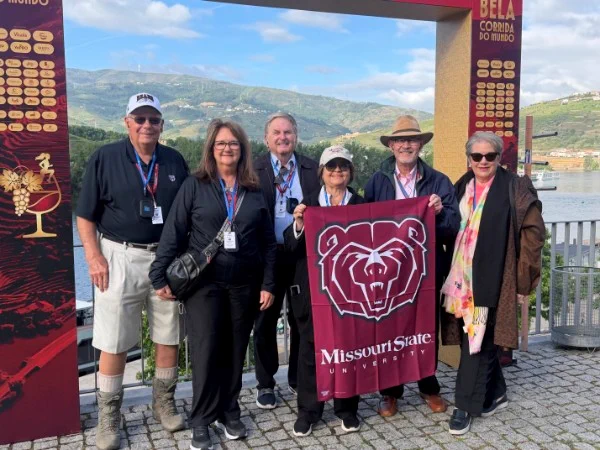 A group of six people is standing on a stone-paved area in front of a scenic river and hills. They are posing under a decorative archway. Two of them are holding a Missouri State University flag. Everyone is wearing casual outdoor attire and lanyards.