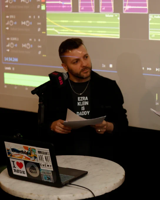 A podcast host presents at an event, holding papers with audio editing software projected in the background. The mood is professional and focused.