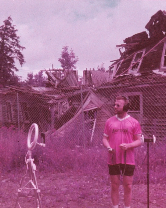A man wearing headphones and a pink shirt stands in front of a dilapidated building. The scene has a vintage, purple-toned look, giving it an artistic and nostalgic feel.