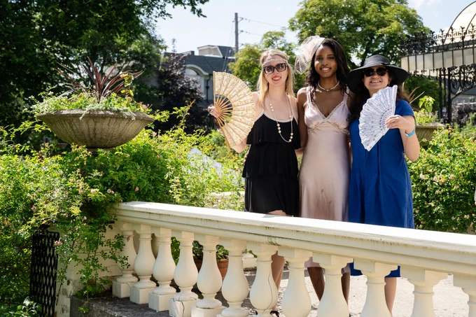 Three women dressed in elegant dresses stand outdoors on a sunny day, each holding a decorative hand fan near a white balustrade.
