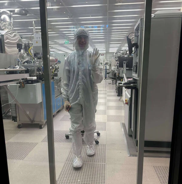 A person in a cleanroom suit is standing behind a glass door in a laboratory filled with advanced scientific equipment. The environment is sterile, with bright lighting and a tiled floor. The person is smiling and waving, holding a phone in the other hand.