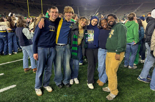 A group of people stand together on a football field, smiling and posing for a photo. The field is filled with others in the background, possibly celebrating an event. Some individuals wear apparel displaying "Notre Dame." The scene is lively and joyful.
