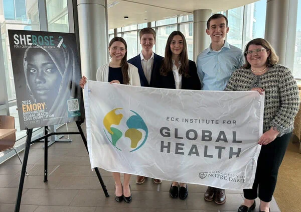 A group of five people stands indoors, holding a banner that reads "Eck Institute for Global Health, University of Notre Dame." Beside them is a poster with the text "SHEROES: 2023 Emory Innovation Case Competition" and a QR code.