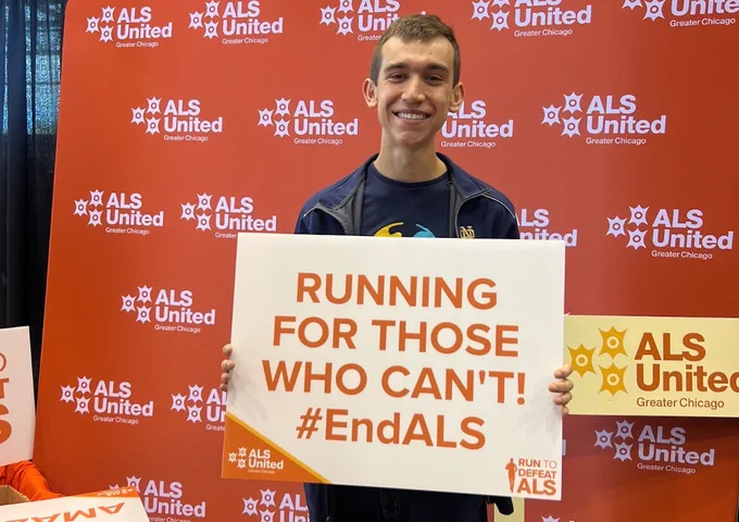 A person smiling while holding a sign that reads, "Running for those who can't! #EndALS" in front of a red backdrop with ALS United Greater Chicago logos.
