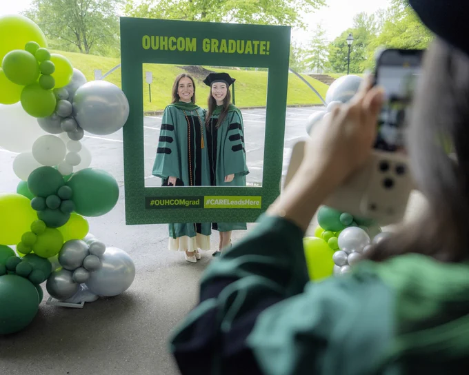 Graduates wearing academic regalia pose within a large green picture frame with "OUHCOM Graduate!" written on top. A person in the foreground takes their picture. The setup is outdoors, with green and white balloon decorations on either side.