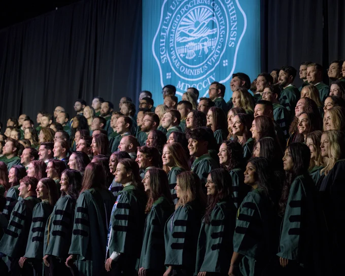 A large group of OHIO Heritage College graduates in green robes stand on a stage, facing forward. 