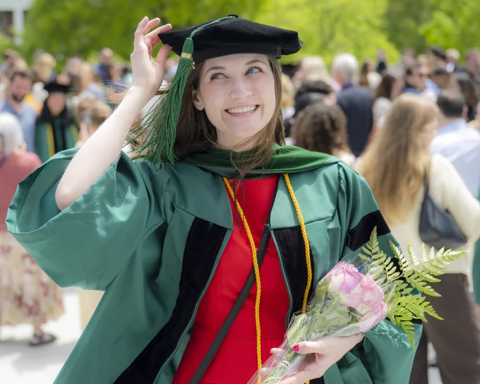 A person in graduation attire, holding a bouquet of pink roses, smiles while adjusting their cap. The setting is outdoors, with a crowd of people and trees in the background.