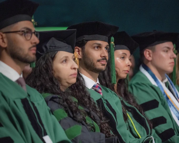 A group of people in graduation attire, including green robes and caps with tassels, sit in a row, focused on a ceremony.