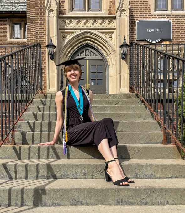 A graduate posed in a cap and sash on the steps of Egan Hall