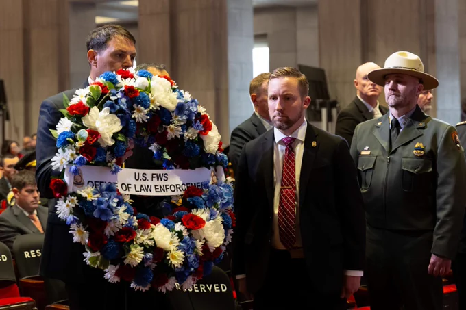 A group of men in formal attire and uniform holding a large wreath with red, white, and blue flowers labeled 'U.S. FWS Office of Law Enforcement'.