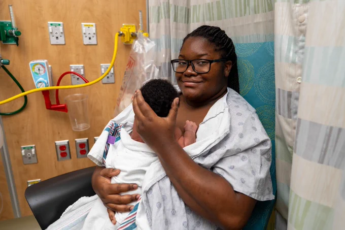 Mother in hospital gown cradling newborn baby wrapped in white blanket while seated in medical room.