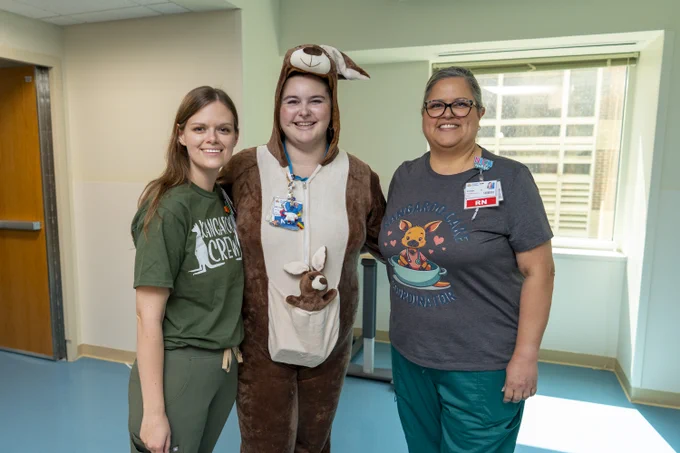 Three people standing indoors, one dressed in a kangaroo costume with a pouch and two others wearing casual clothing with animal-themed shirts.