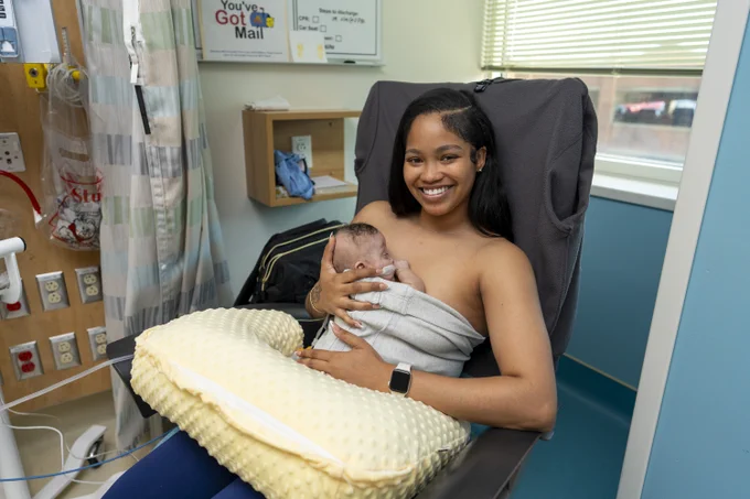 Mother sitting in a hospital chair breastfeeding her newborn wrapped in a blanket with a nursing pillow on her lap.