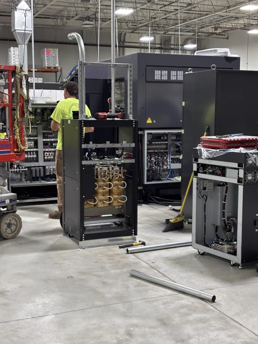 Worker in hi-vis shirt wiring large industrial electrical cabinets with open panels and tools nearby.