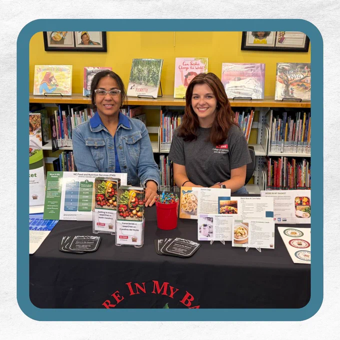Two women at a library table sitting behind a booth with food and nutrition materials about the More In My Basket program.