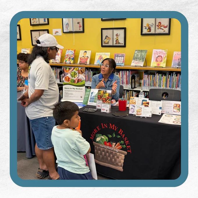 Community event table for "More In My Basket" program with a woman talking to a man and child, surrounded by books and informational materials.