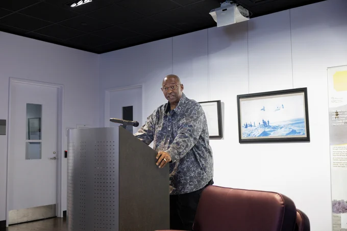 A person stands at a podium giving a speech in a modern room. The walls display framed art.