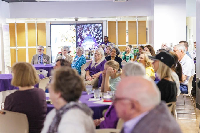 A group of people sits in a brightly lit room, attentively listening to a presentation. The room features purple tablecloths and large windows in the background.