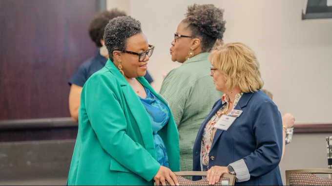 Two people are engaged in conversation in a conference setting. One is wearing a green blazer, and the other is in a blue blazer with a name tag. Others are visible in the background, standing and talking. The atmosphere appears professional and social.