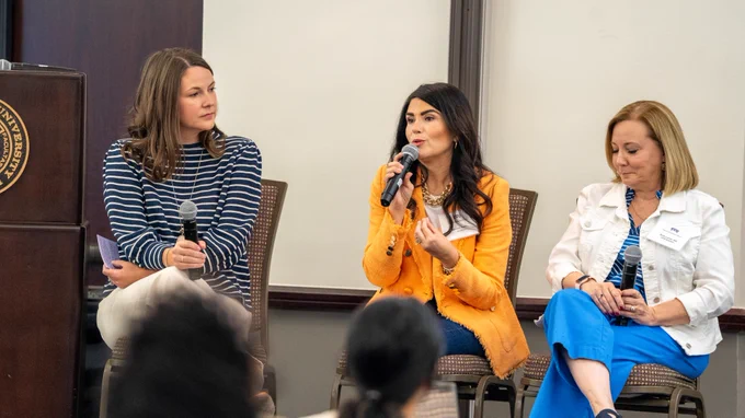 A panel of three people seated on chairs, holding microphones, engaged in discussion. The setting appears to be a university event, as indicated by a visible part of a university emblem on a podium. They are in a conference room with an audience.
