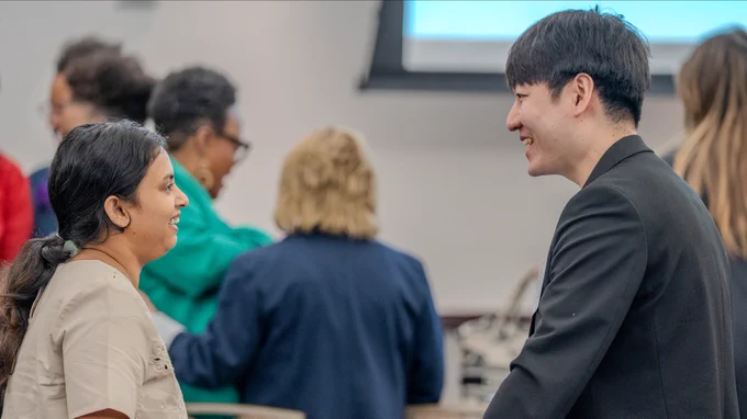 Two people are smiling and talking to each other in a conference room setting. Other attendees are conversing in the background.