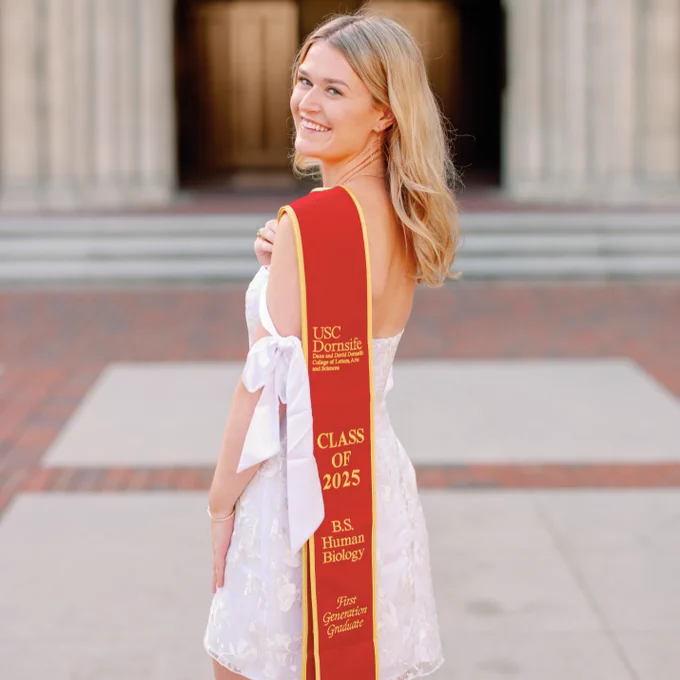 A person in a light dress stands smiling, holding a red sash over their shoulder. The sash reads "USC Dornslife, Class of 2025, B.S. Human Biology, First Generation Graduate." They stand in an outdoor setting with a blurred building in the background.