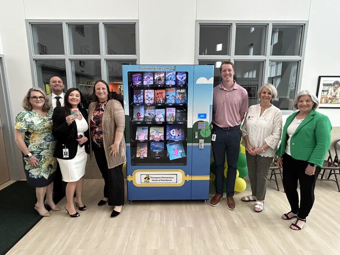A group of people standing in front of a vending machine displaying various books. The people appear to be in casual business attire and are smiling.
