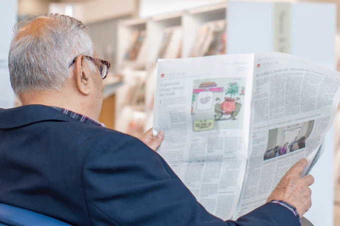 A person with glasses is seated, reading a newspaper. Books are visible on shelves in the background.