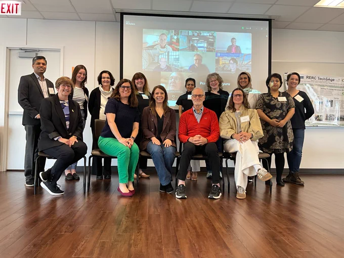 Group photo of TechSAge team members and board members. In-person attendees are posing in front of projector showing attendees on Zoom.