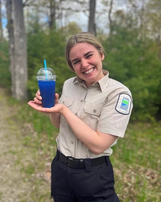 Smiling person in an Ontario Parks uniform holding up a blue slushie drink. There is a background of green trees.