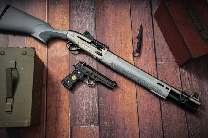 A tactical shotgun and a Beretta pistol are displayed on a wooden floor. Nearby are two storage boxes and a small knife.